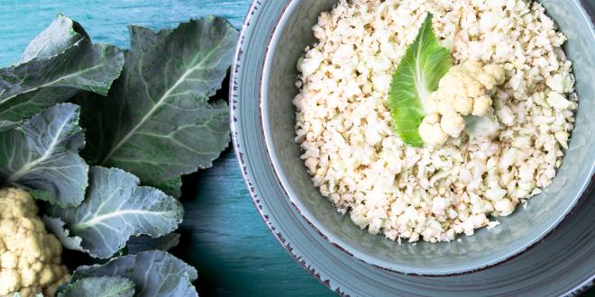 Cauliflower rice in a blue bowl on a blue table next to a head of cauliflower with leaves.
