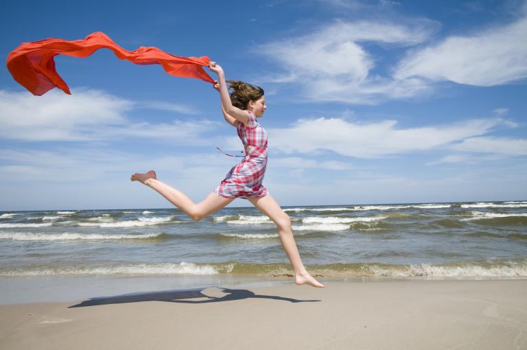 A teenage girl runs along a beach trailing a red cloth behind her.